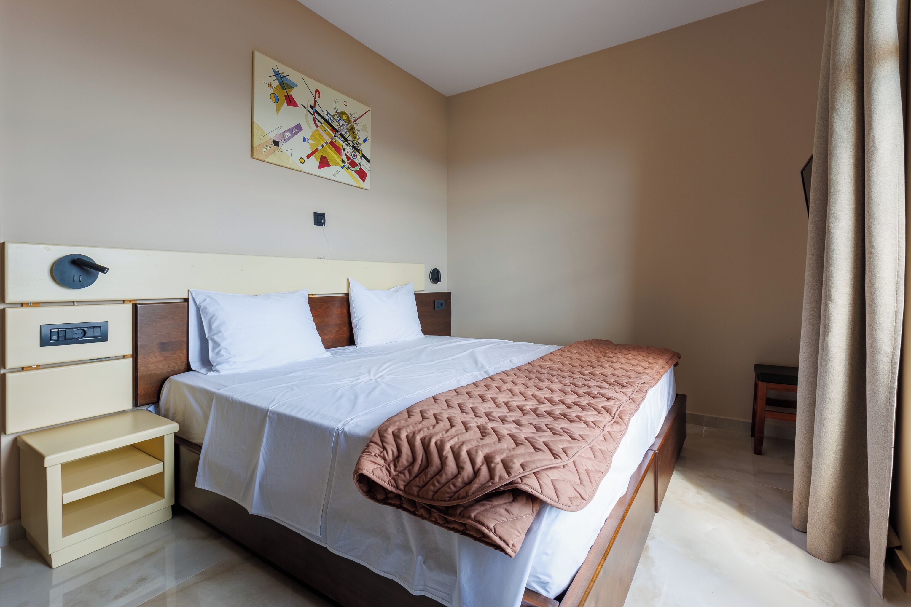 A hotel room with a bed, bedside tables, a nightstand, a painting on the wall, and a window with curtains.