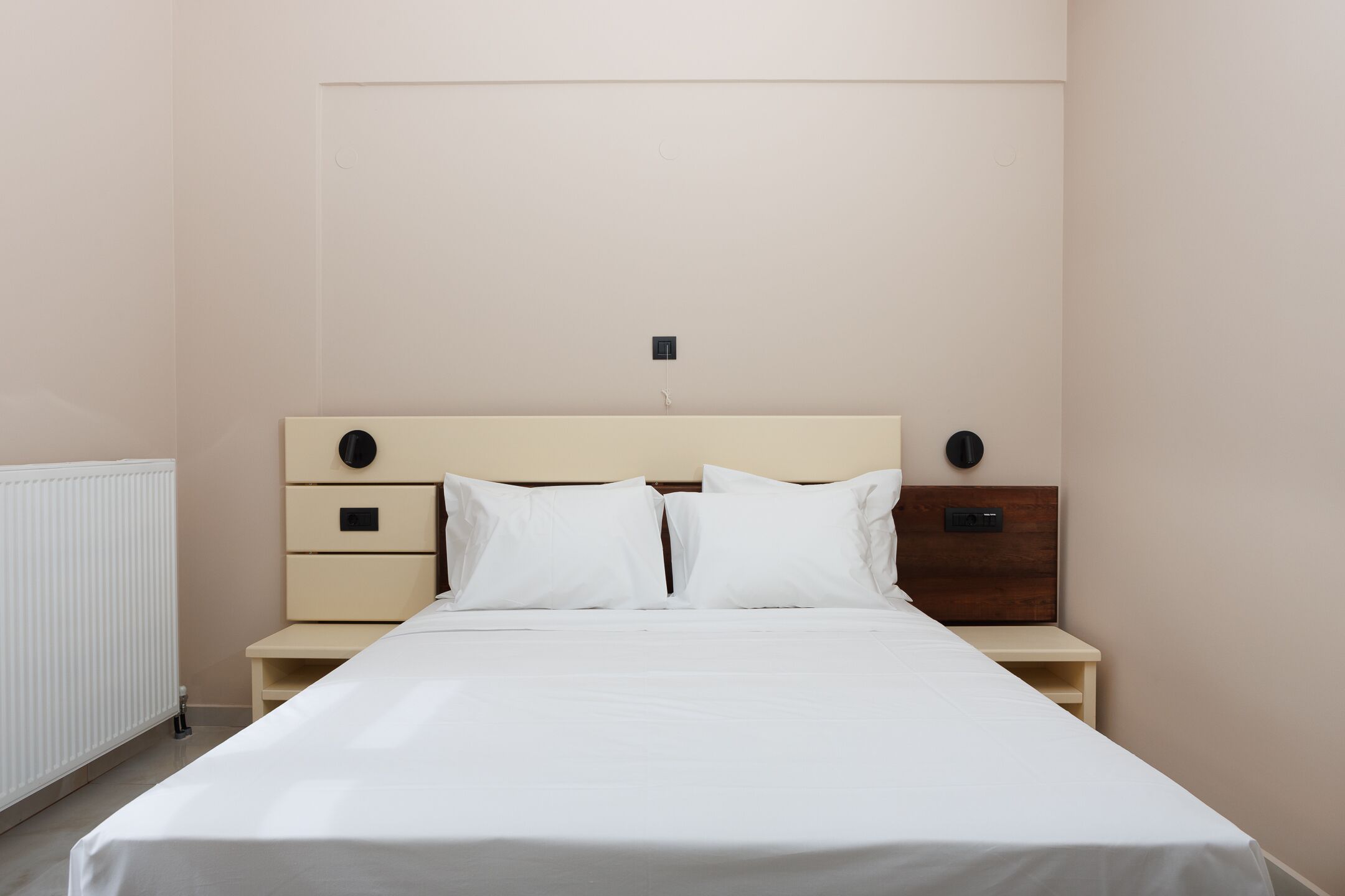 A neatly made bed in a hotel room with a simple headboard, two bedside tables, and a wall-mounted light fixture.