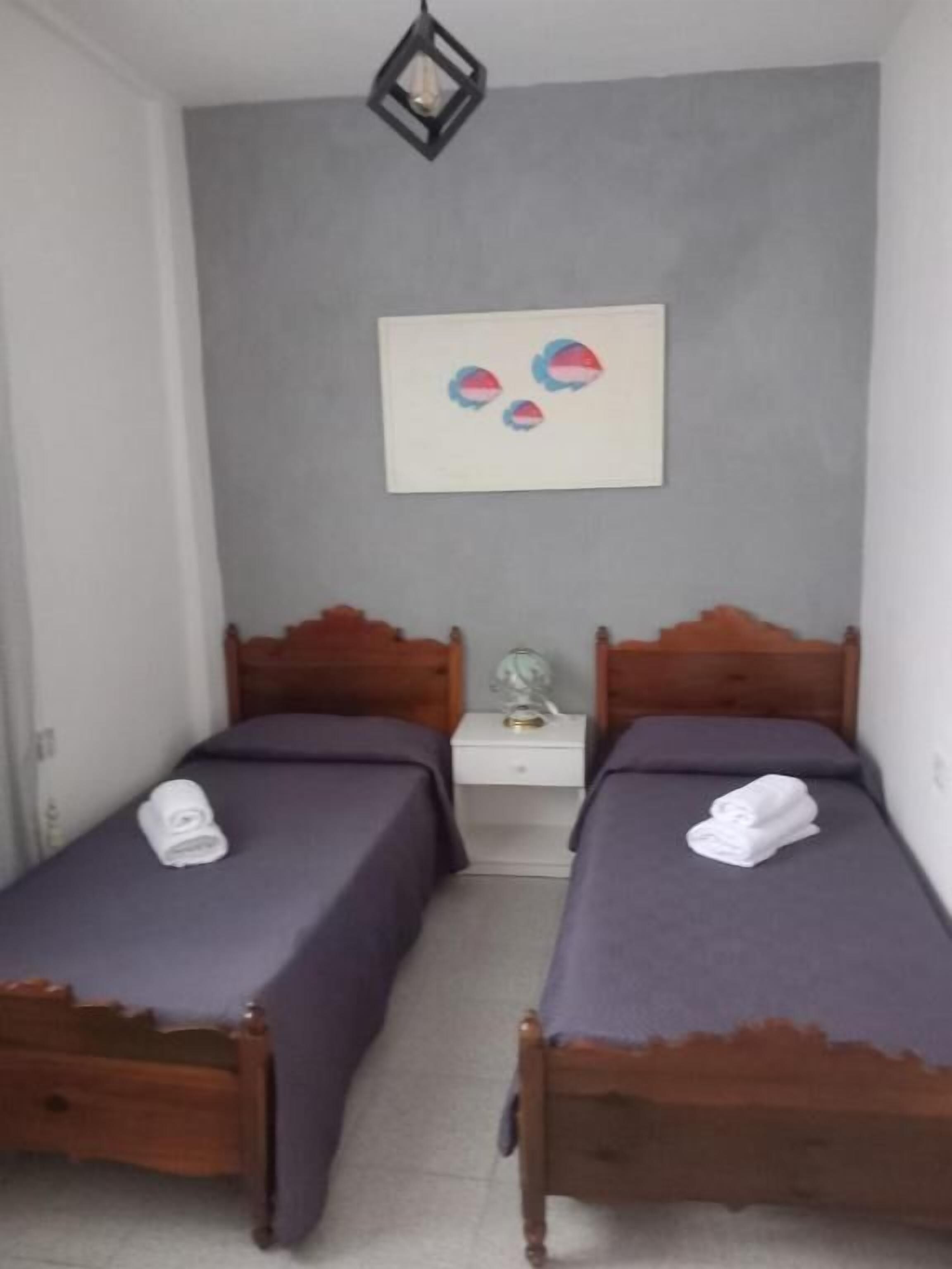 A room with two wooden beds, each with a purple bedspread and a rolled white towel on the bed. There is a white nightstand between the beds with a lamp on it. Above the beds, there is a framed artwork on the wall.