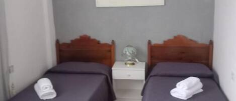 A room with two wooden beds, each with a purple bedspread and a rolled white towel on the bed. There is a white nightstand between the beds with a lamp on it. Above the beds, there is a framed artwork on the wall.