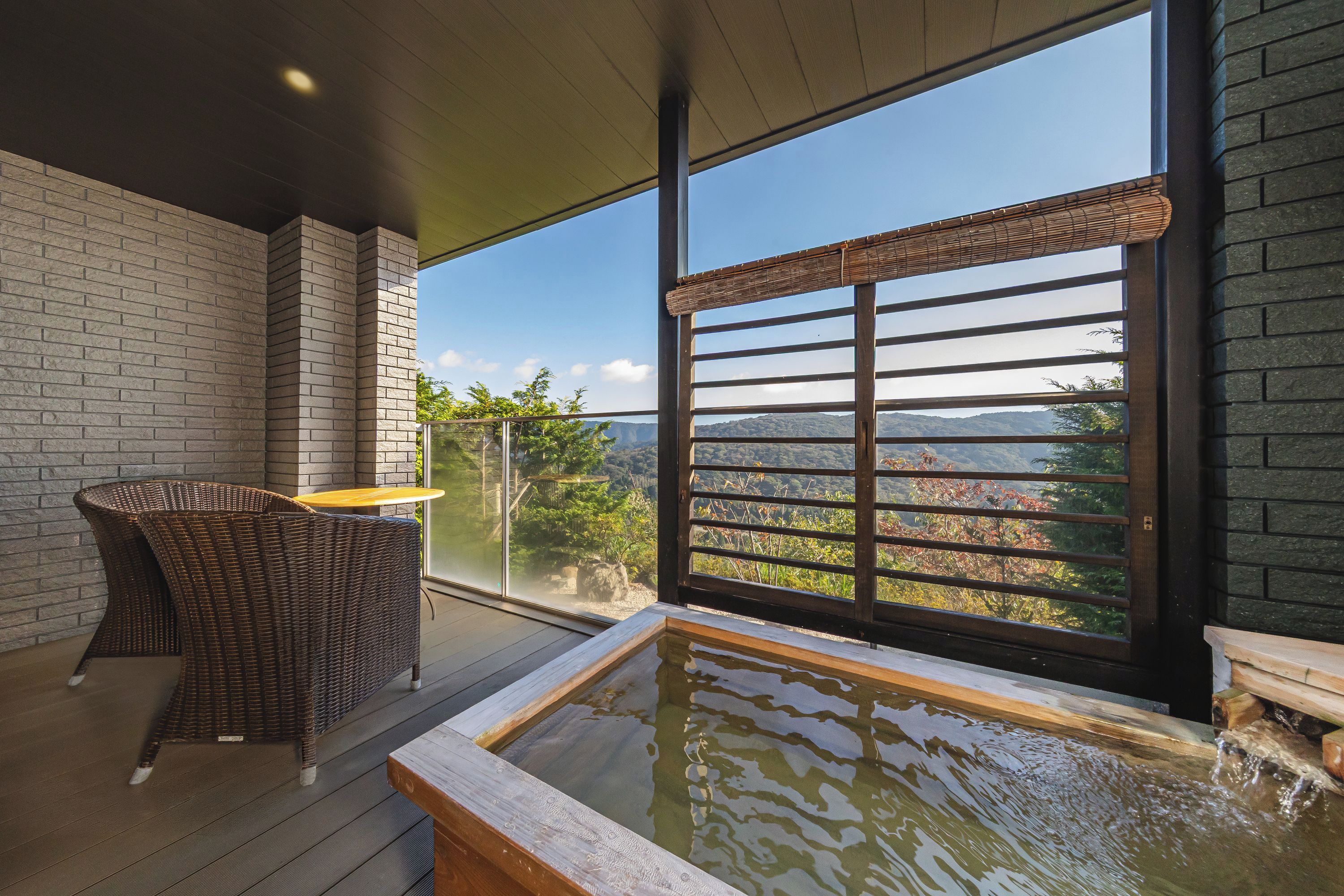 A balcony with a view of a mountainous landscape, a wooden table, and a wicker chair.