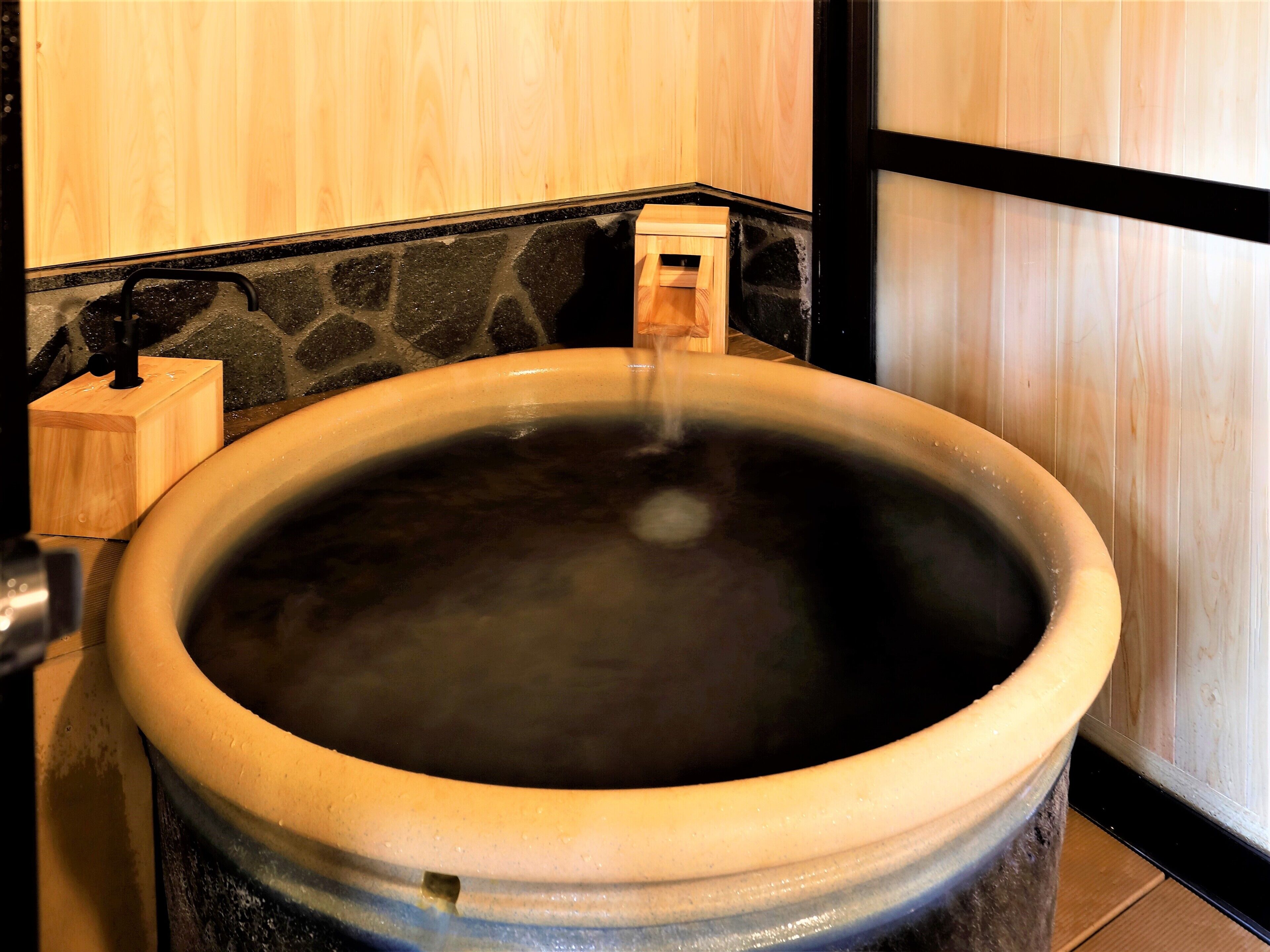 A large ceramic hot tub with a wooden stand and a faucet, set against a wooden wall.