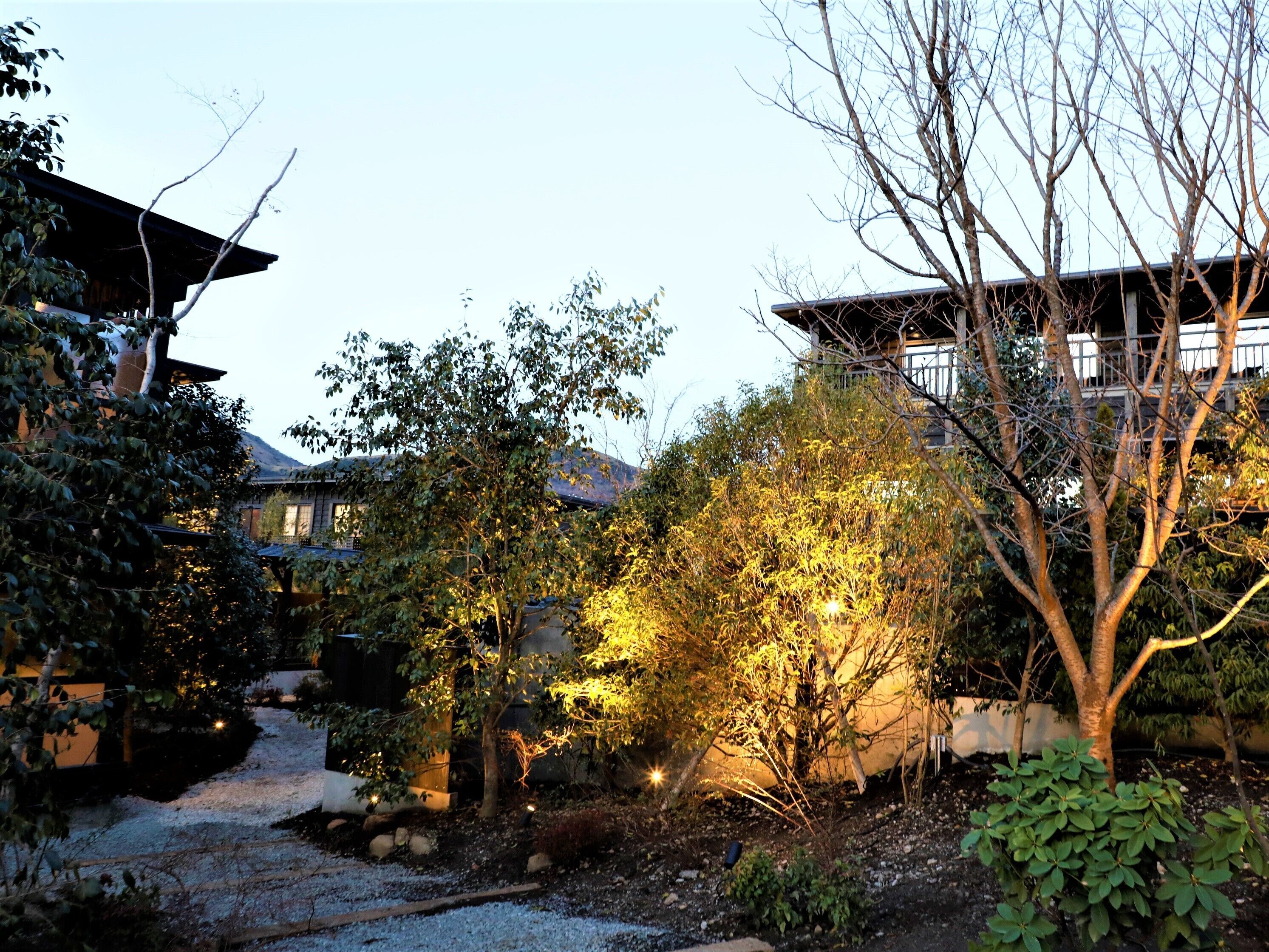A traditional Japanese-style garden with a gravel path, various trees, and a building with a sloped roof.