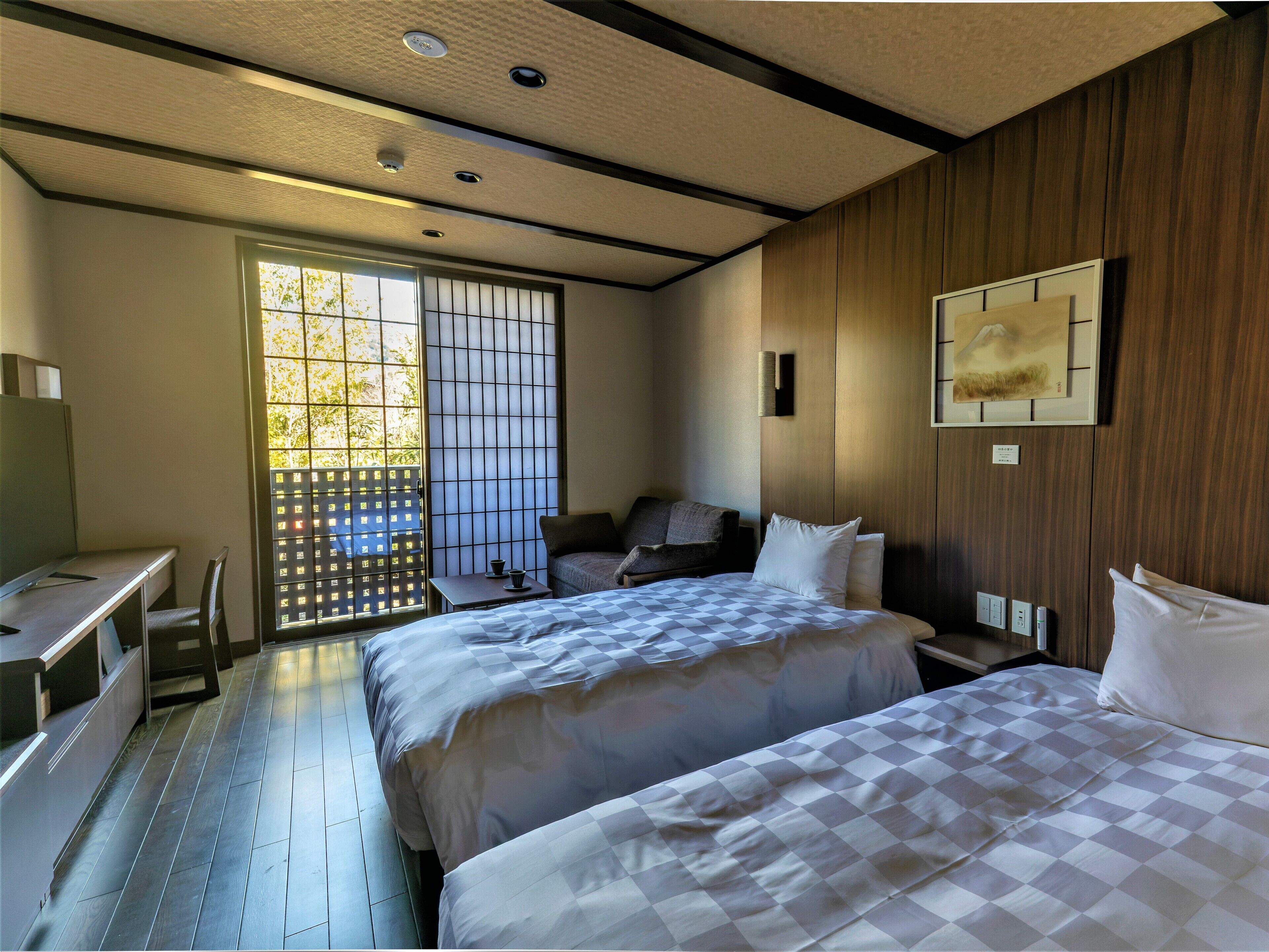 A hotel room with two beds, a desk, a chair, and a sliding door to a balcony.