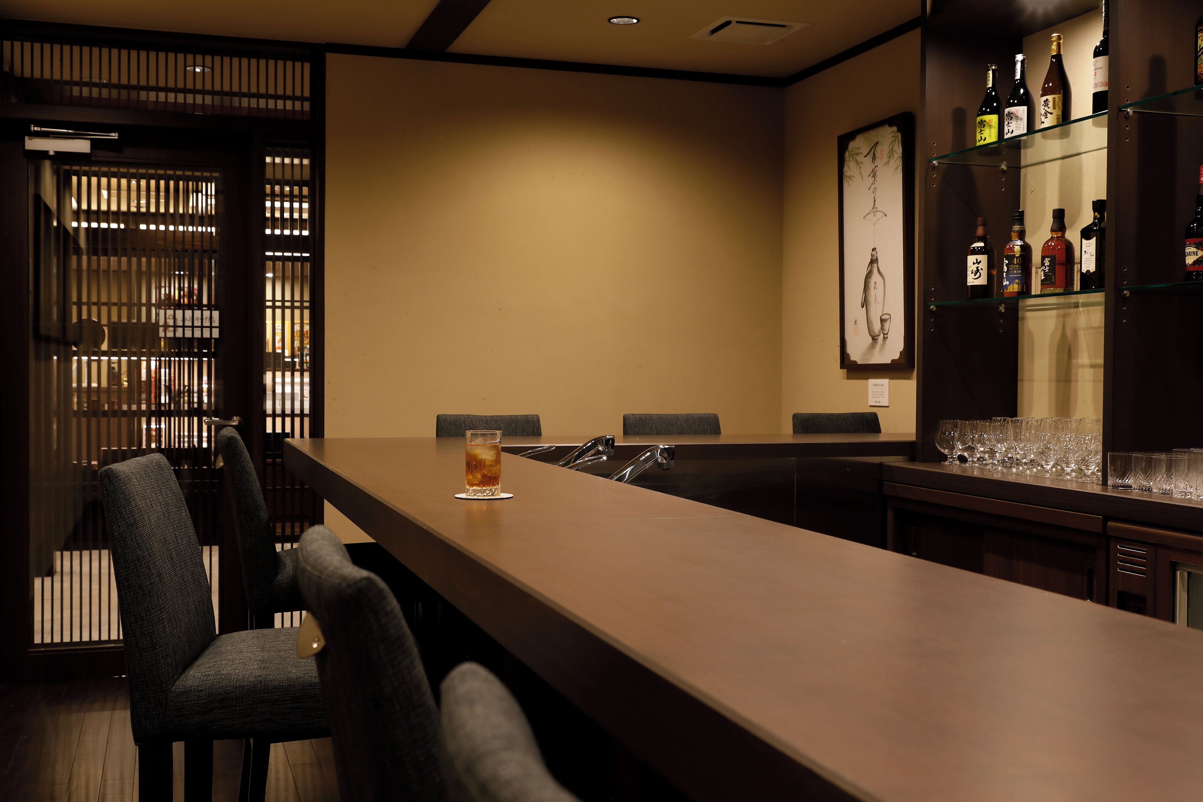 A bar area with a long counter, several chairs, and a glass of beer. There are shelves with bottles and glasses behind the counter.
