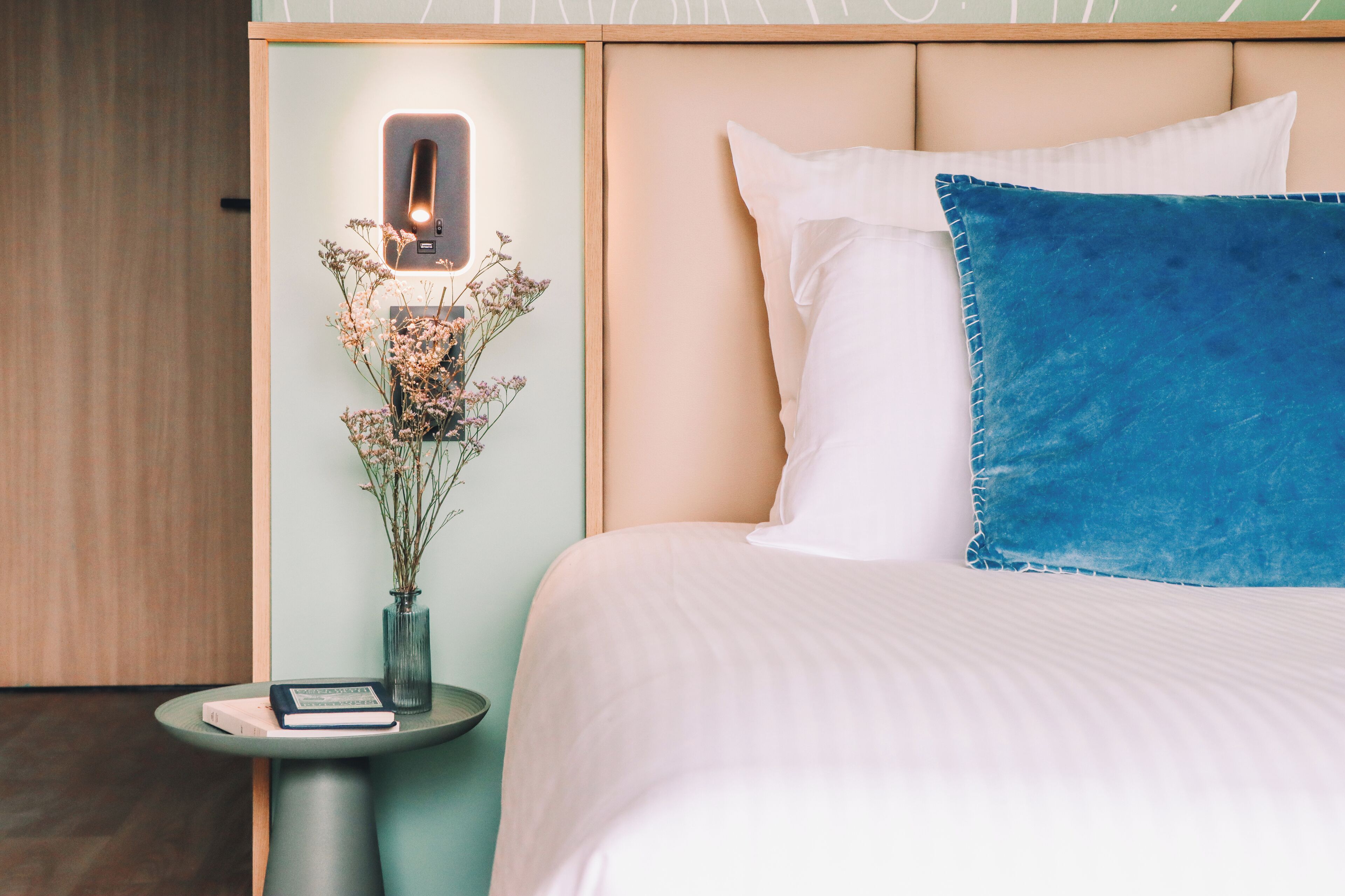 A neatly made bed with white and blue pillows, a bedside table with a book and a vase of dried flowers, and a wall-mounted light fixture.