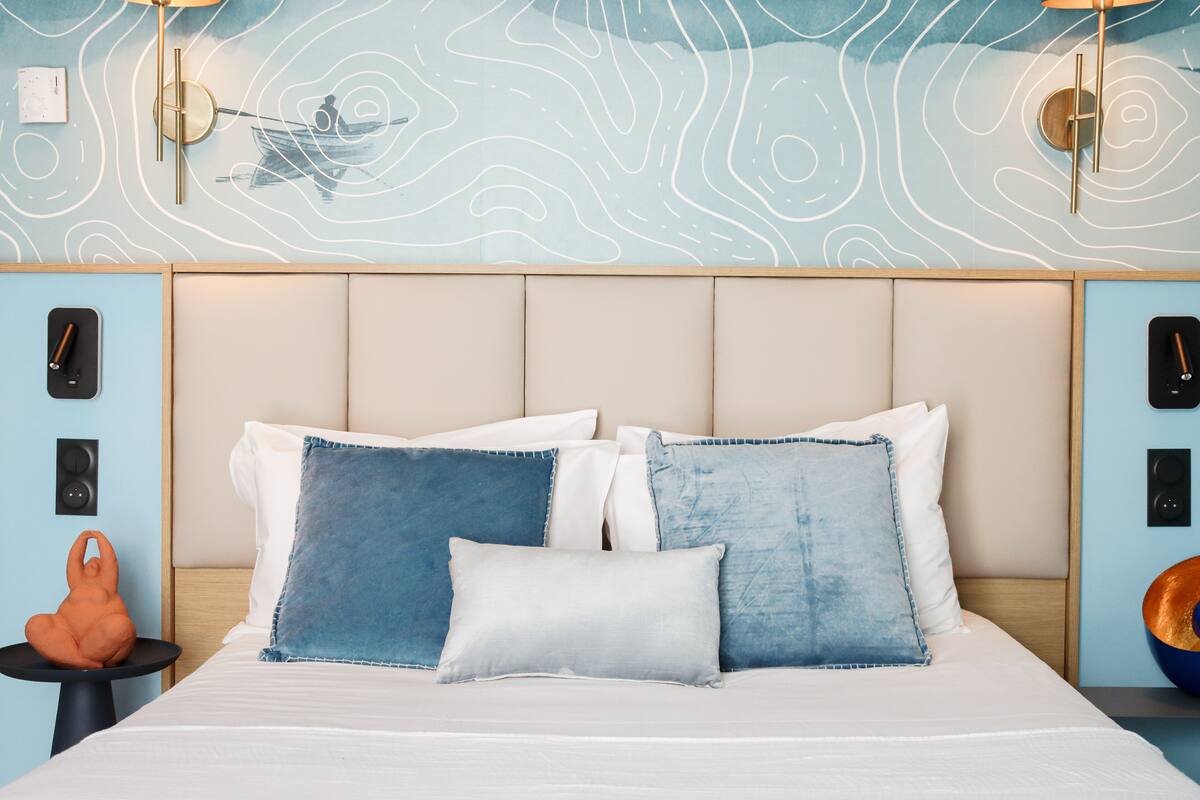 Um quarto com cama, almofadas, uma parede com padrão de ondas e uma pequena mesa com um objeto decorativo.