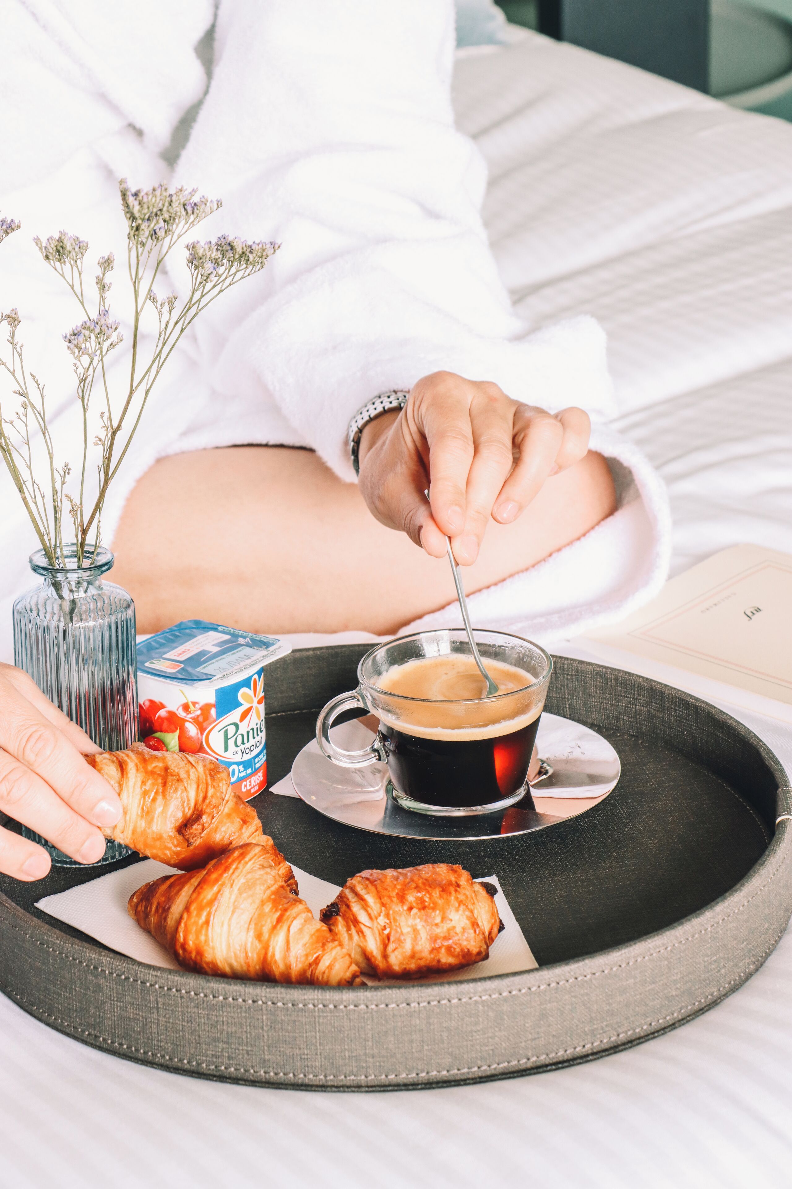 En bakke med en croissant, et glas kaffe og en karton yoghurt på en seng.