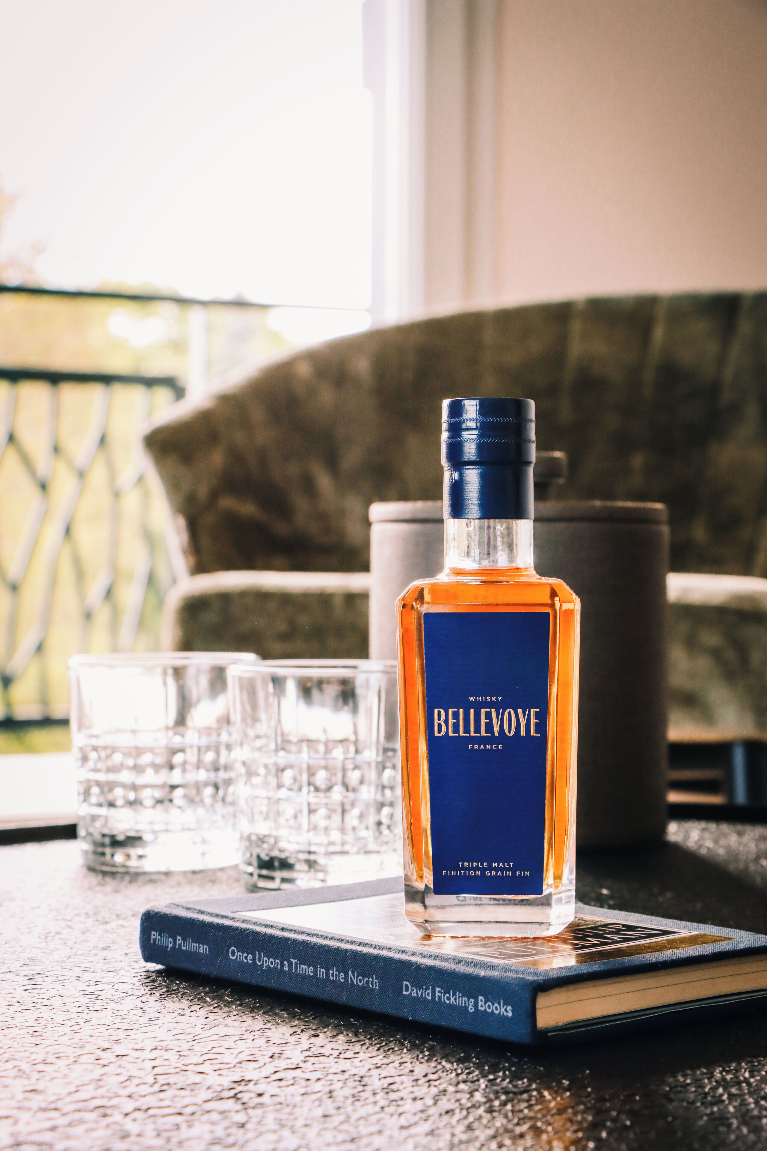 Een fles whisky op een boek getiteld 'Once Upon a Time in the North' van Philip Pullman, met glazen en een stoel op de achtergrond.