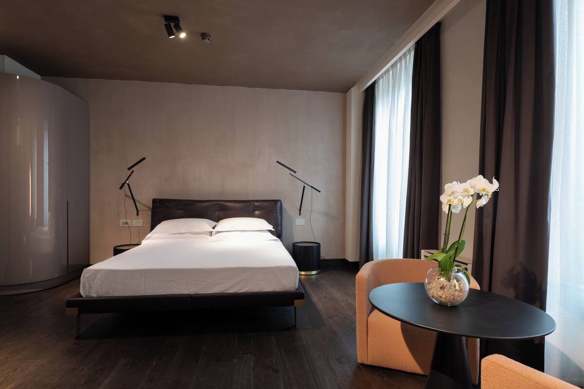 A modern hotel room with a large bed, a bedside table with a vase of flowers, and a view of the city through large windows.