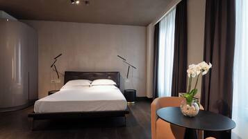 Ett modernt hotellrum med en stor säng, ett sängbord med en vas med blommor och utsikt över staden genom stora fönster.