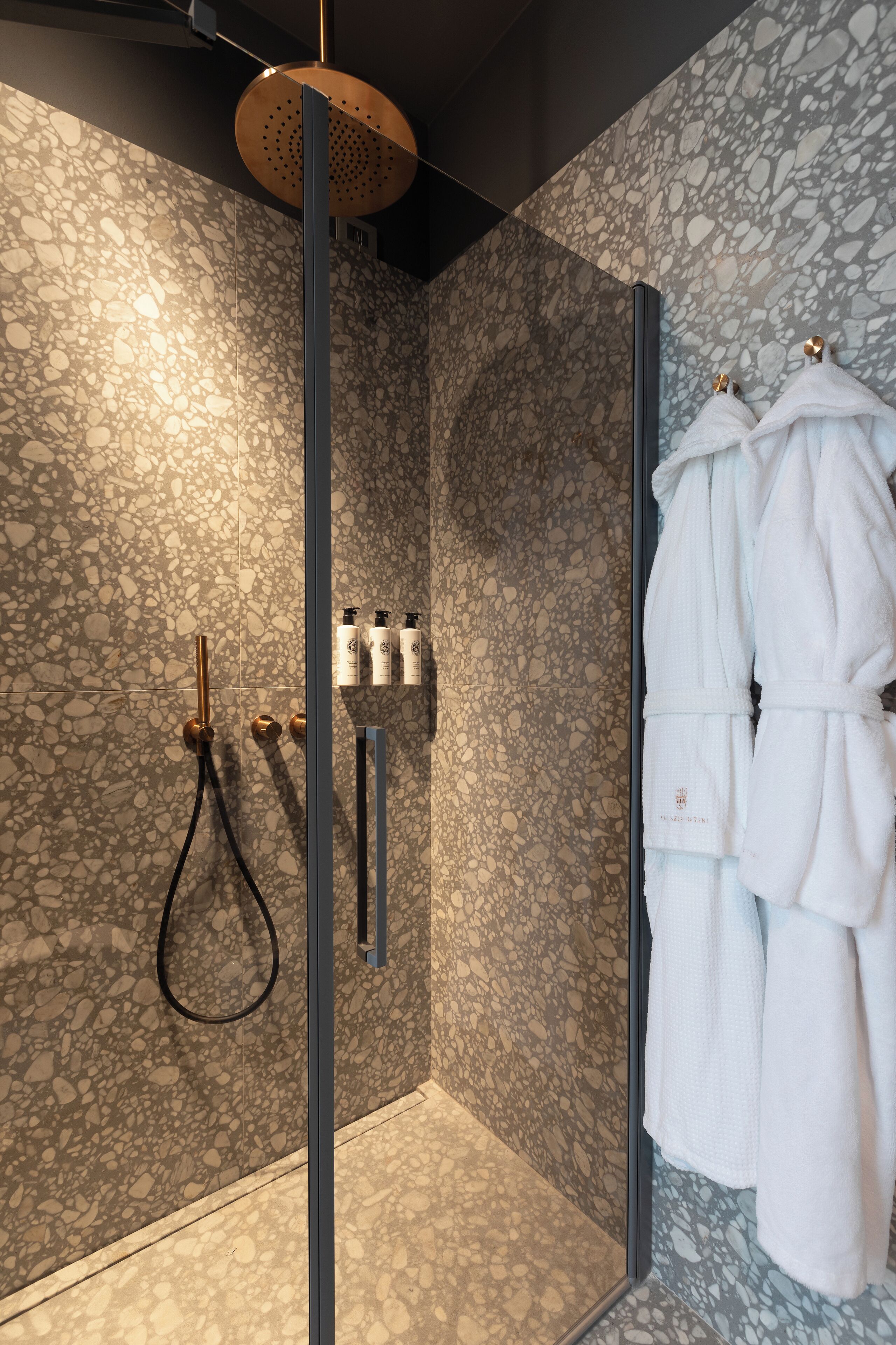 Un baño moderno con pared de textura de guijarros, ducha con cerramiento de vidrio y batas blancas colgadas en ganchos.