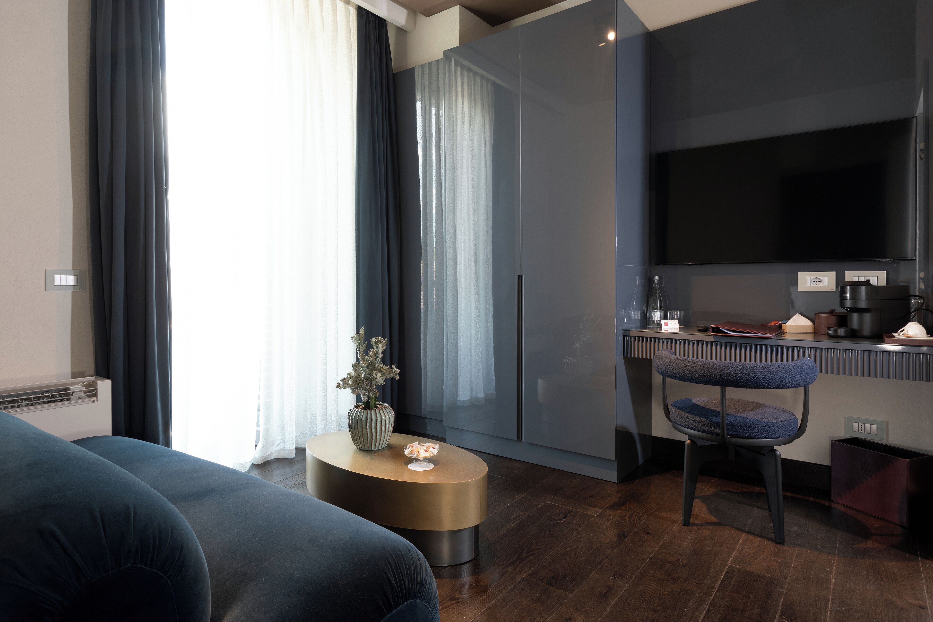 A modern hotel room with a dark blue sofa, a round wooden side table with a vase, a chair, a flat-screen TV, and a kitchenette area.