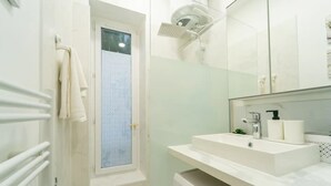 Une salle de bain moderne avec une douche à l’italienne en verre, un lavabo blanc avec un miroir au-dessus, et un porte-serviettes sur la gauche.