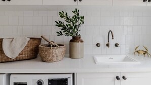 A kitchen with white cabinets, a sink, a Bosch washing machine, wicker baskets, a potted plant, and a decorative crab. - Port Cottage on the Great Ocean Road (Port Campbell)