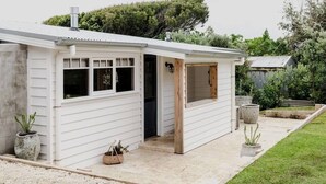 A small, single-story house with a white exterior, a wooden door, and a window. There are potted plants outside and a gravel driveway. - Port Cottage on the Great Ocean Road (Port Campbell)