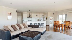 Ein modernes Wohnzimmer mit Ecksofa, Hocker, Essbereich mit Holzkästen, Küche mit weißen Schränken, Pendelleuchten und einem großen Fenster.