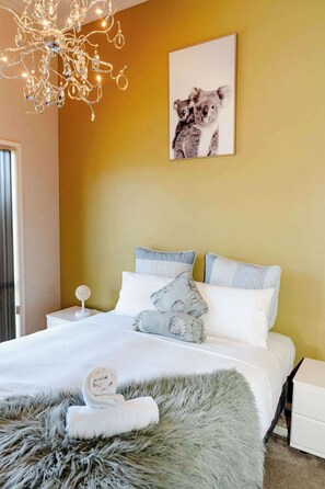 A bedroom with a bed, pillows, a fluffy rug, a nightstand, a lamp, and a wall-mounted picture of koalas.