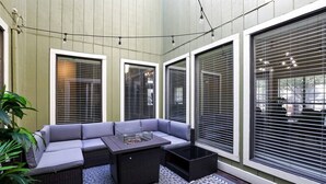 Een modern buitenverblijf met een grijze bank en een salontafel met glazen blad, omringd door grote ramen met luxaflex.