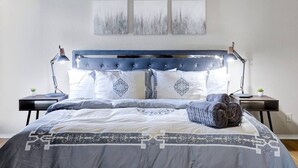 Een slaapkamer met een gebordureerd hoofdeinde, een groot bed met beddengoed met een patroon, twee nachtkastjes met lampen en drie aan de muur bevestigde kunstwerken.