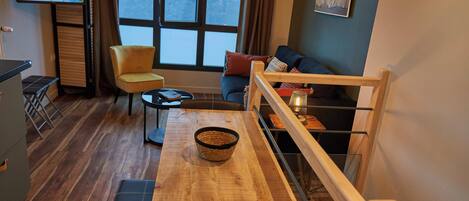 A modern living room with a wooden table, a comfortable sofa, a TV, and a view of the water outside.