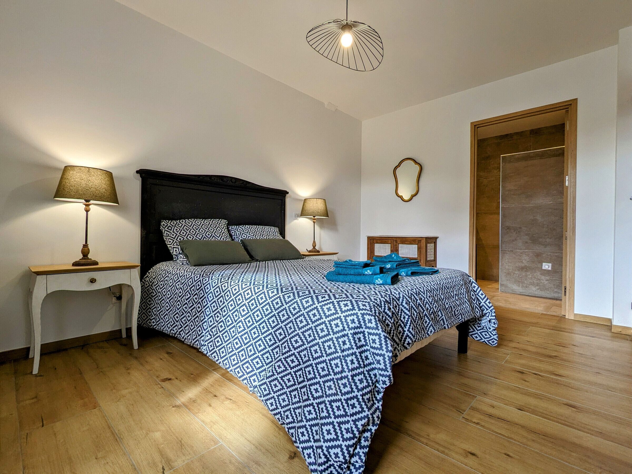 A bedroom with a patterned bedspread, two bedside lamps, a wooden nightstand, a mirror, and a wooden door.