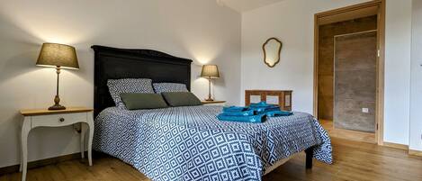 A bedroom with a patterned bedspread, two bedside lamps, a wooden nightstand, a mirror, and a wooden door.