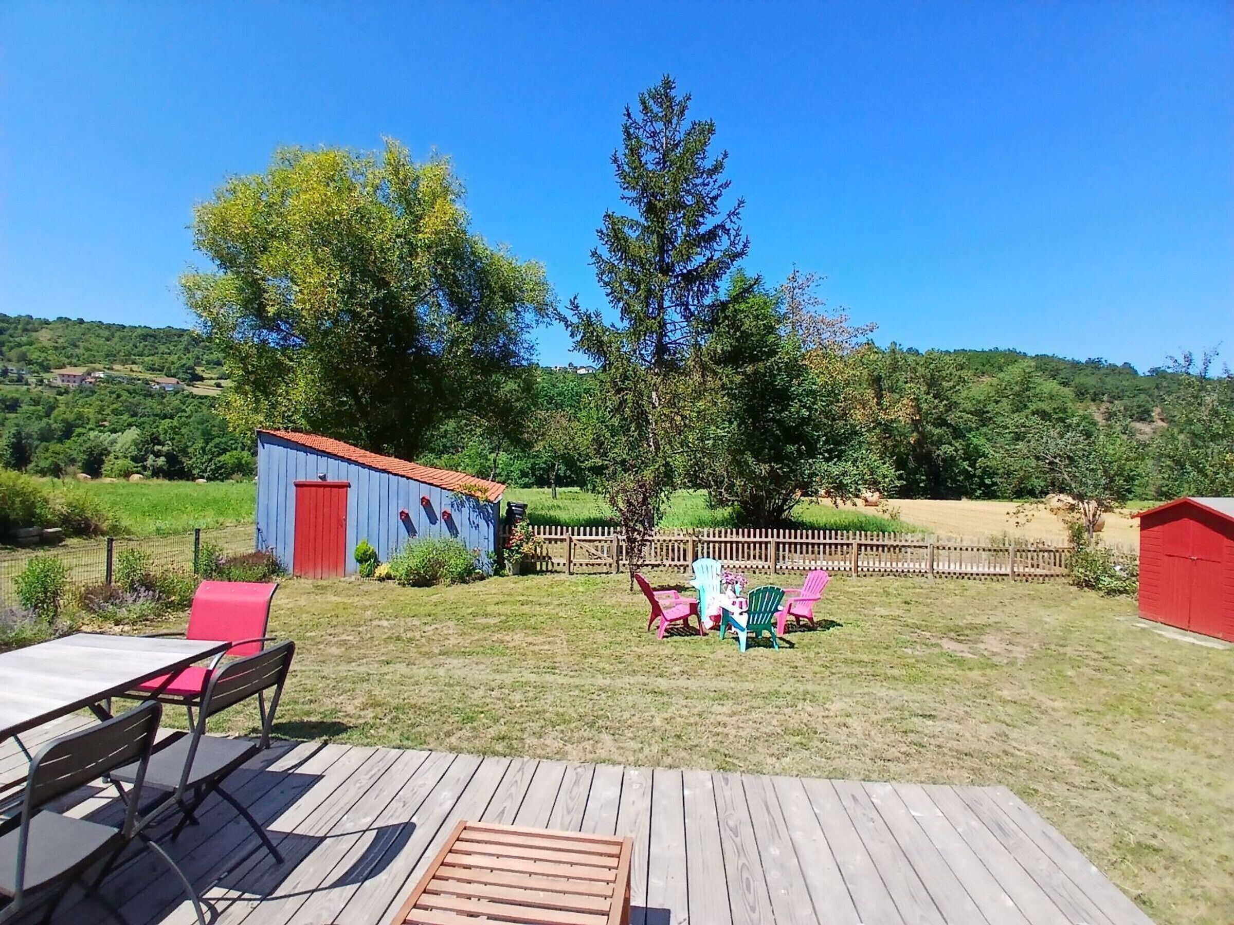 A wooden deck with outdoor furniture, a blue shed, and a red barn in a rural setting.