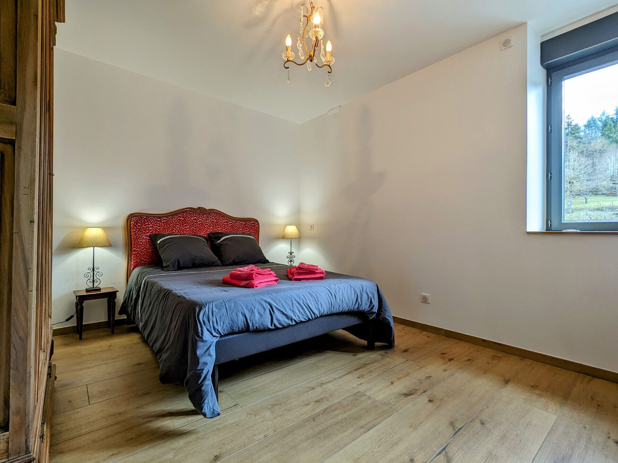 A bedroom with a bed, two bedside lamps, a chandelier, a window, and a wooden floor.