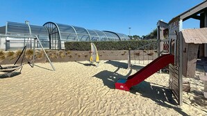 Een zandstrand met een schommel, een rode glijbaan en een kasstructuur op de achtergrond.