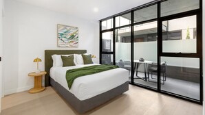A modern bedroom with a large bed, a side table, a green throw blanket, and a view of the outside balcony through a glass door. - Fitzroy North Apartments by Urban Rest (Clifton Hill)