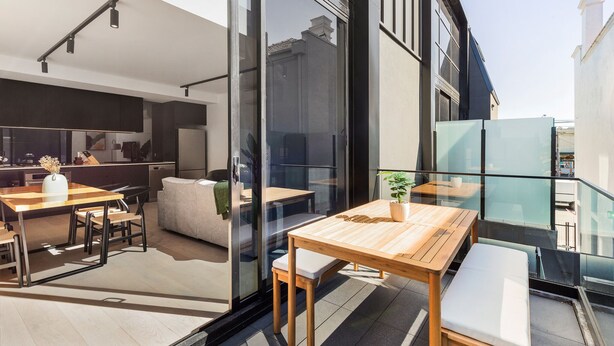 A modern outdoor dining area with a wooden table and benches, a glass railing, and a view of the cityscape. - Fitzroy North Apartments by Urban Rest (Clifton Hill)