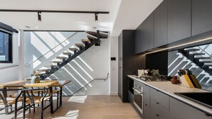 A modern kitchen with a built-in stove, a dining area with a wooden table and chairs, and a staircase with a glass railing. - Fitzroy North Apartments by Urban Rest (Clifton Hill)