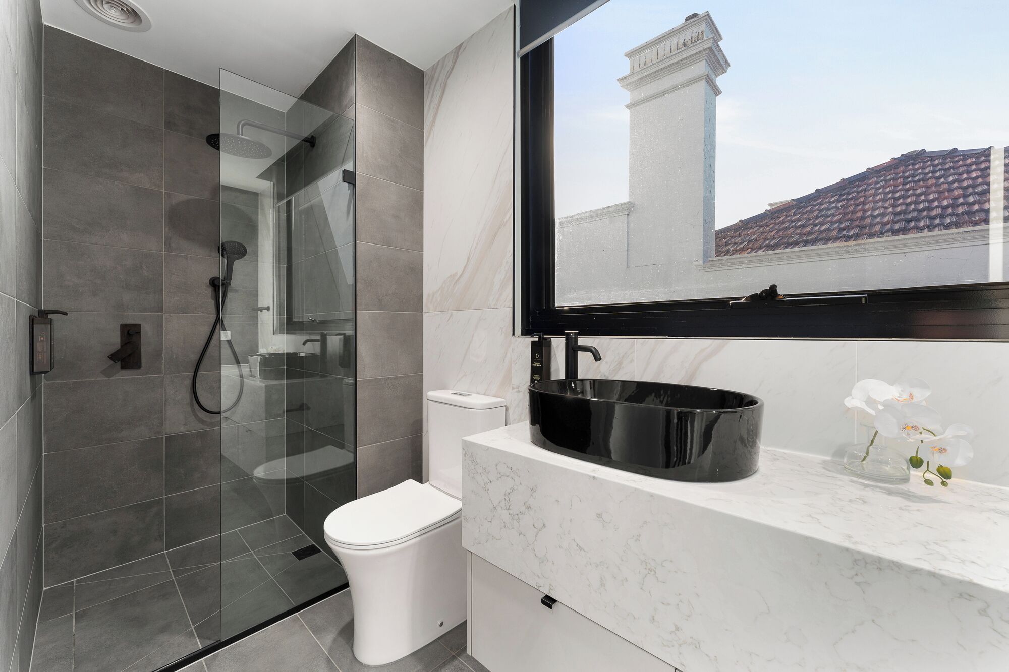 Une salle de bain moderne avec une douche à parois de verre, des toilettes blanches, un lavabo noir et un comptoir en marbre.