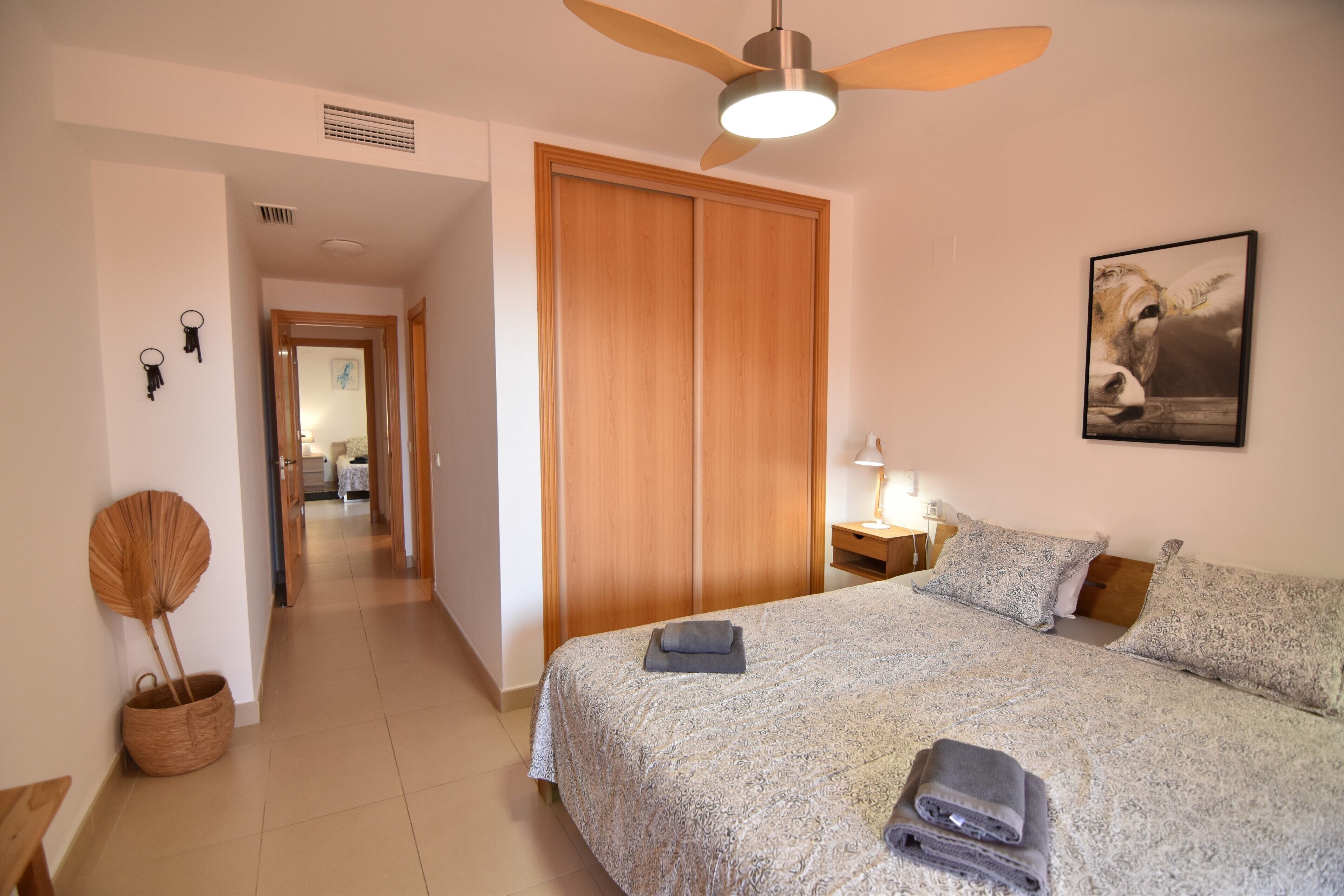 Una camera d'hotel con un letto, un armadio in legno, una ventola a soffitto, una lampada e un quadro appeso al muro.