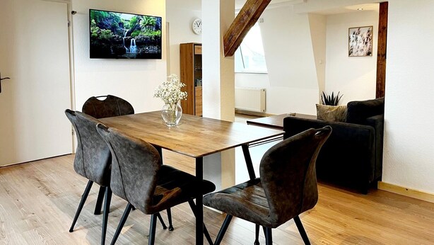 A modern living room with a wooden dining table, dark leather chairs, a TV on the wall, and a staircase leading to an upper level. - "Lindenblatt" 3-room apartment, 2 bathrooms, Büsum (4km), parking lot, North Sea (Oesterdeichstrich)