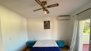 A bedroom with a bed, a ceiling fan, an air conditioning unit, and a window with curtains.