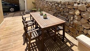 An outdoor patio with a wooden table and chairs, surrounded by a stone wall and a grassy slope.