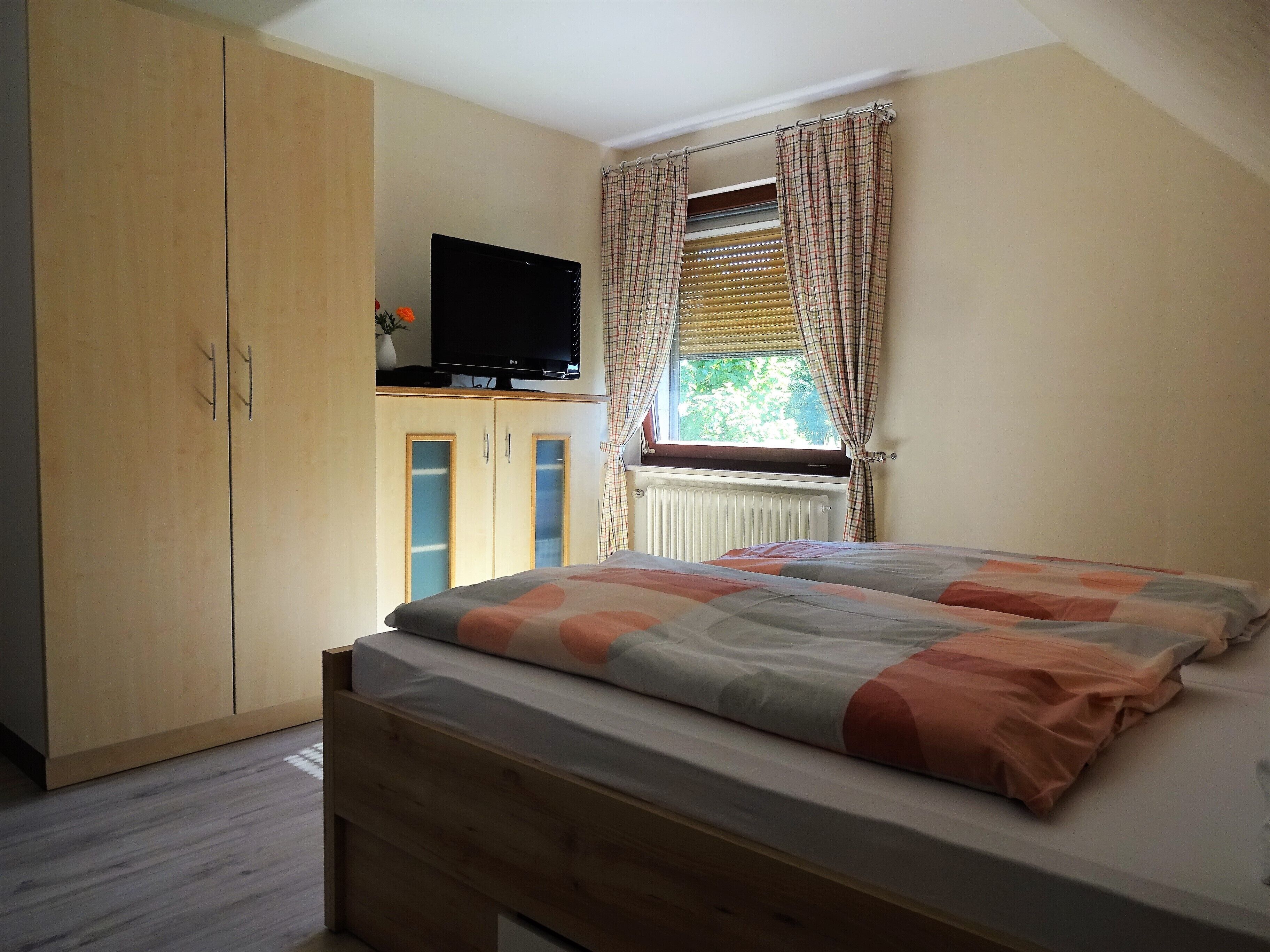 Une chambre à coucher avec un lit, une armoire, un téléviseur posé sur un meuble et une fenêtre avec des rideaux.