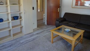 Sala de estar com sofá, mesa de centro, estante de livros e porta para outro cômodo.