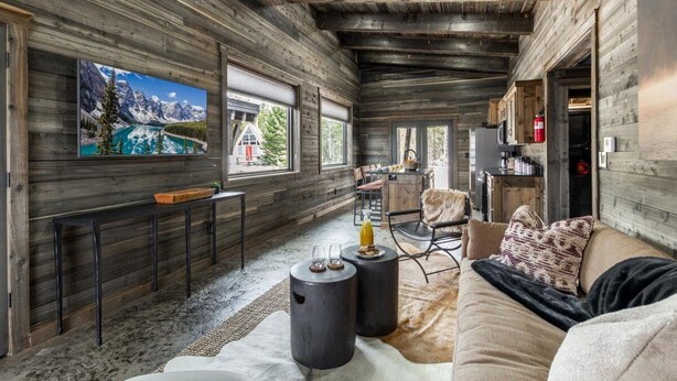 A rustic cabin interior with a wooden interior, a television mounted on the wall, a sofa, and a dining area with chairs. - Pet Friendly Home!- Mayfly Meadow | Cozy Cabin | World-Class Fishing | Fire Pit | BBQ (Island Park)