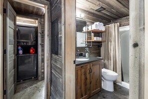 A compact bathroom with a built-in sink, toilet, and shower area. It features wooden cabinetry and a small refrigerator with cleaning supplies.