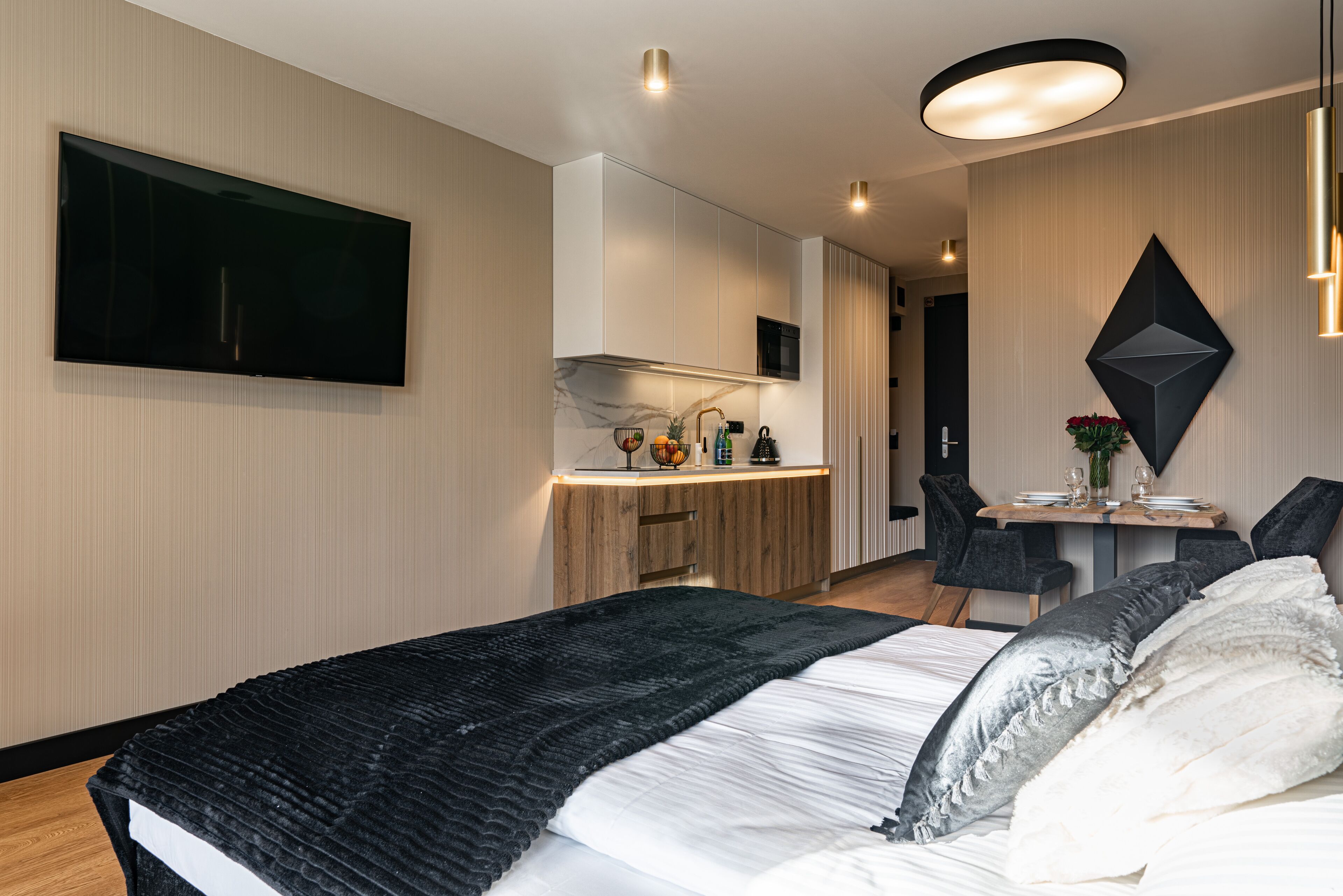 A modern hotel room with a large bed, a flat-screen TV mounted on the wall, a small kitchen area with a microwave, and a dining table with chairs.