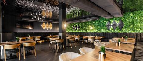 A modern restaurant with wooden tables and chairs, a green wall with plants, and geometric wall decorations.