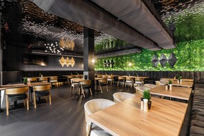 Ein modernes Restaurant mit Holztischen und -stühlen, einer grünen Wand mit Pflanzen und geometrischen Wanddekorationen.