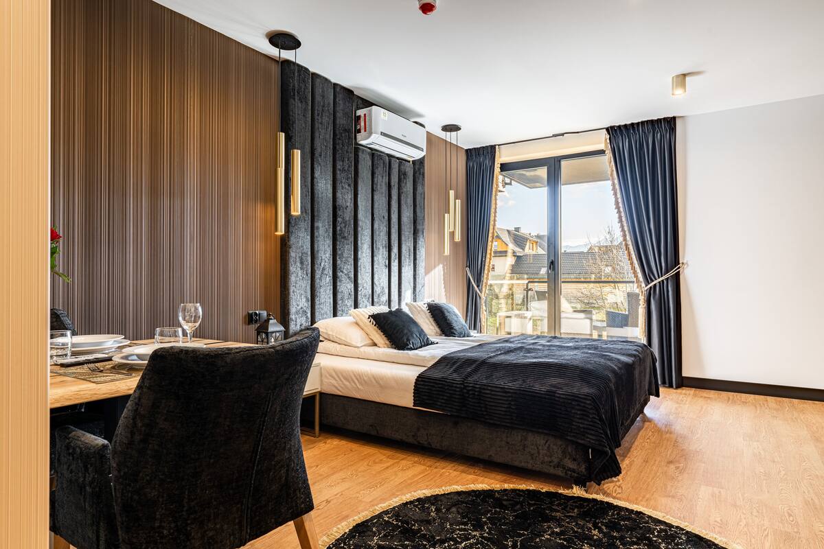 A modern bedroom with a bed, a dining table with chairs, and a large window with curtains.
