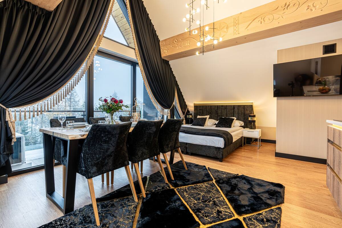 A modern bedroom with a large bed, a dining area with chairs, and a view through a window.