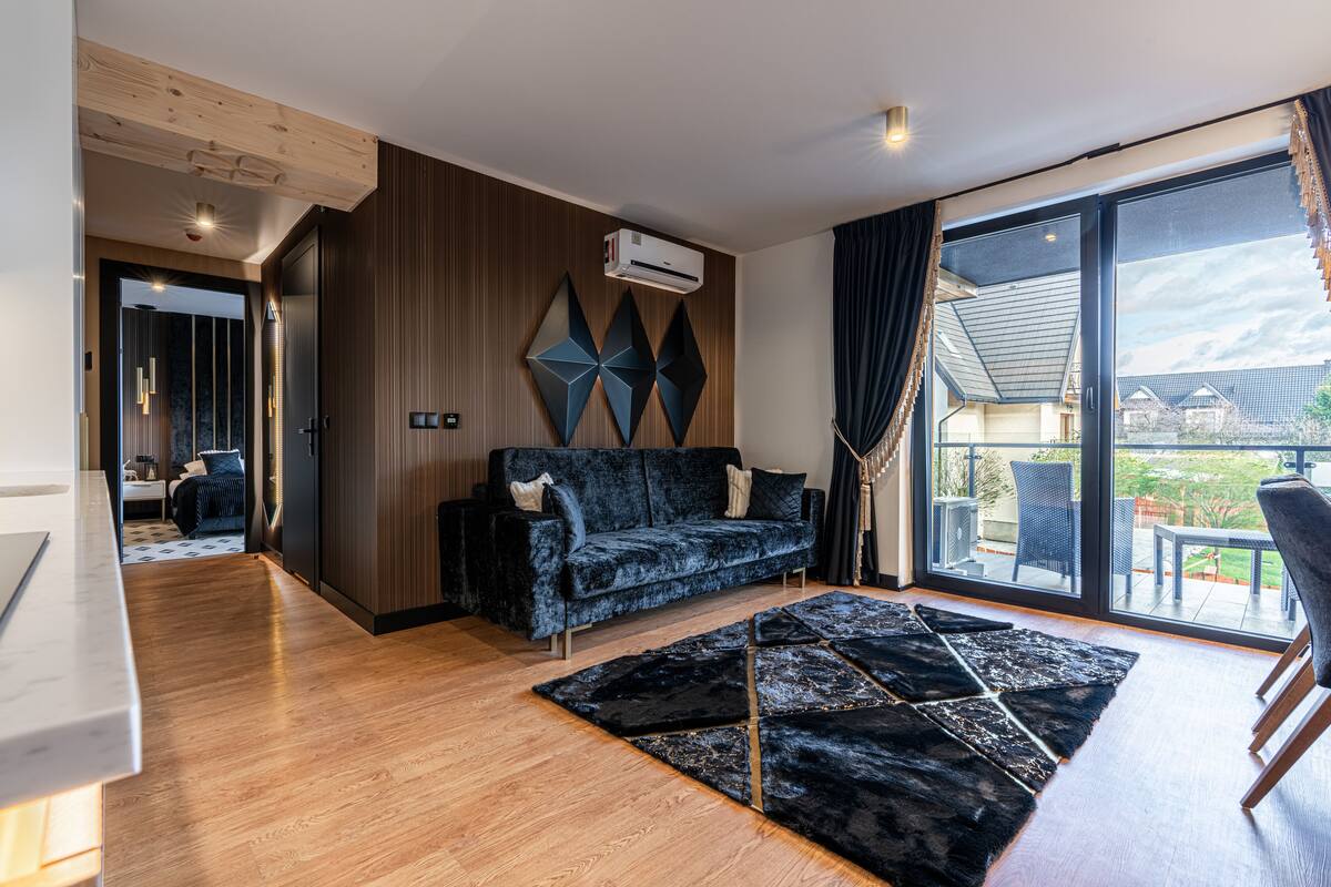 A modern living room with a dark blue sofa, a geometric rug, and a large window offering a view of the outdoors.