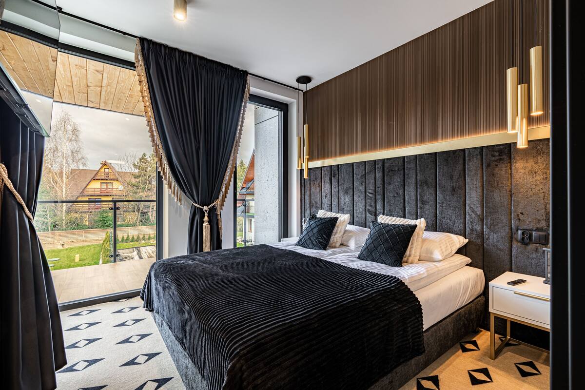 A modern bedroom with a large bed, a nightstand, and a view of a house through a glass door.