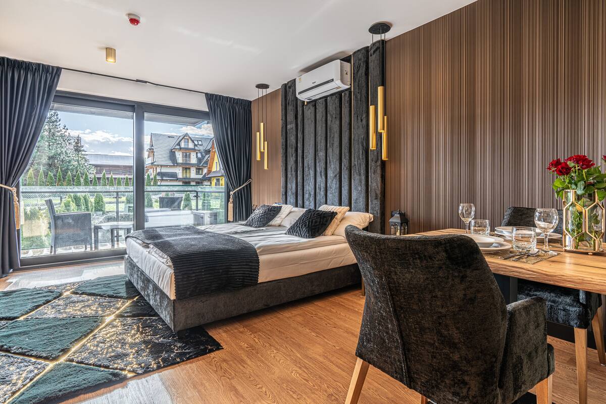 A modern bedroom with a large bed, a dining table set for two, and a view of a house through the window.
